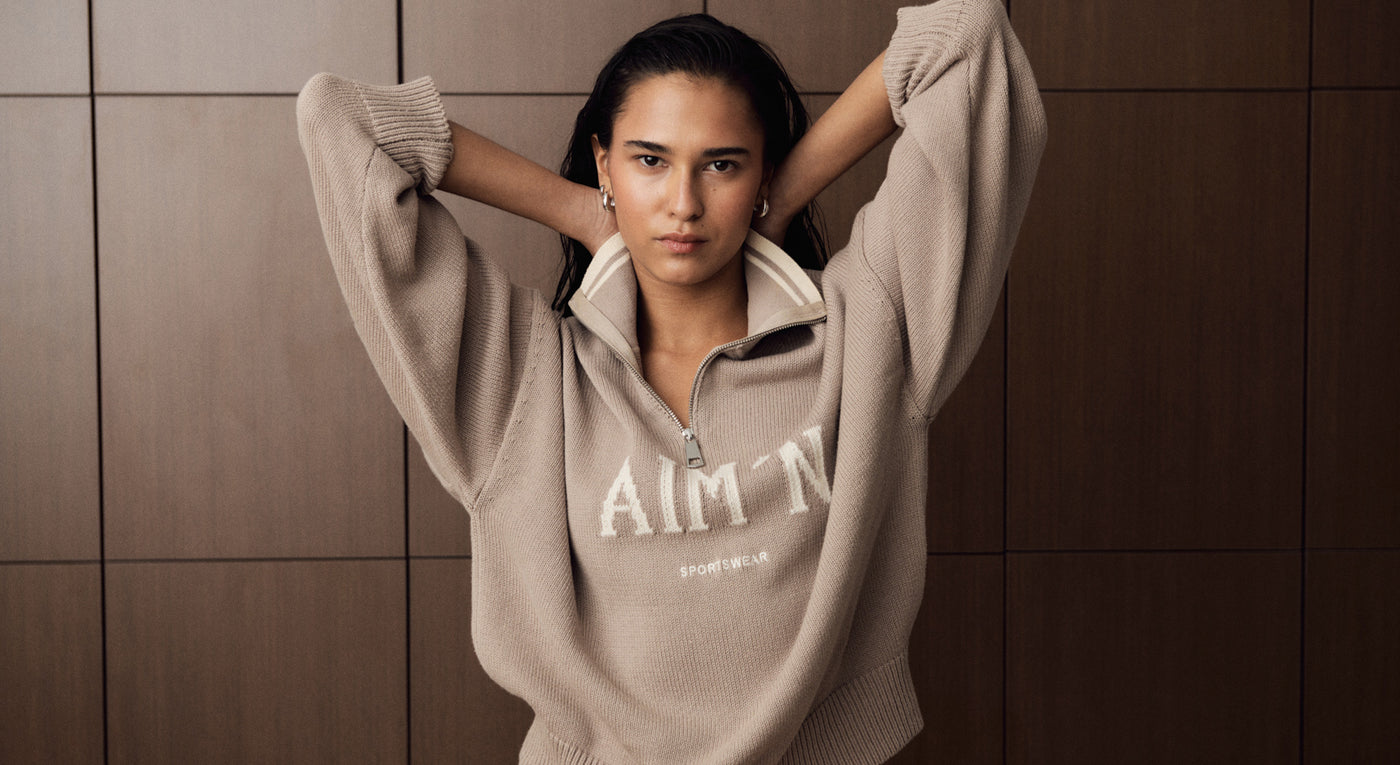 Das Model trägt den Dark Clay College Knitted Half Zip. Sie posiert mit den Händen hinter dem Kopf vor einer Holzpaneelwand.