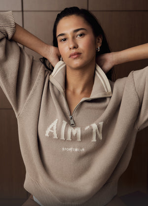 Das Model trägt den Dark Clay College Knitted Half Zip. Sie posiert mit den Händen hinter dem Kopf vor einer Holzpaneelwand.
